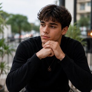 A young man in a black long-sleeve shirt sits outdoors with his hands clasped under his chin, looking off to the side.