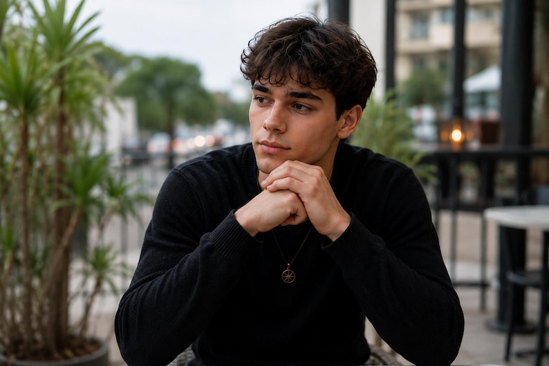 A young man in a black long-sleeve shirt sits outdoors with his hands clasped under his chin, looking off to the side.