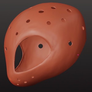 May include: A terracotta-colored protective headgear piece featuring a smooth, rounded exterior shell with a large, central oval-shaped opening for the face and multiple small, circular ventilation holes distributed across the top and sides.
