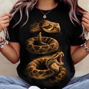 May include: A black t-shirt featuring a graphic print of three rattlesnakes in various poses. The top snake is shown from the head, the middle snake is coiled, and the bottom snake is coiled with its mouth open, displaying sharp fangs.