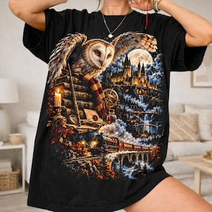Peut inclure: Un t-shirt noir avec une impression graphique détaillée représentant une effraie des clochers perchée sur une pile de livres, une baguette, une bougie allumée et une écharpe. Le fond montre un château sur une falaise, une pleine lune et un train.