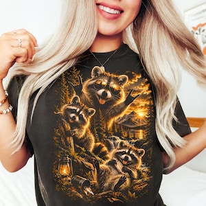 Puede incluir: Una camiseta negra con un estampado gráfico de tres mapaches en un entorno forestal al atardecer. Un mapache lleva gafas de sol y hace un gesto con la mano de rock and roll. La escena incluye una linterna brillante, pinos, montañas y un lago.