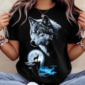 May include: A black t-shirt featuring a large, detailed graphic of a wolf's head in shades of white and blue. Below the wolf, a smaller silhouette of a wolf stands on a rocky cliff against a bright full moon, with blue light illuminating the base.