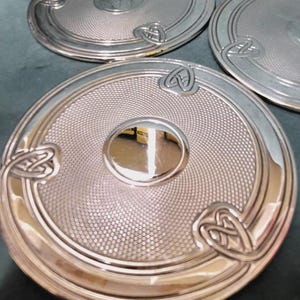 Könnte beinhalten: Drei kreisförmige Untersetzer aus silberfarbenem Metall mit einer glatten, reflektierenden Scheibe in der Mitte, umgeben von einem punktierten Texturmuster und einem äußeren Rand mit drei erhabenen, symmetrischen Knotenmotiven.