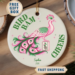 May include: A round white ceramic ornament featuring a pink and green peacock illustration, the text BIRD BAM and CHEERS, and the personalised name Sophia with the year 2026. The ornament includes a gold hanging string at the top centre.