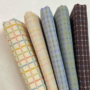 May include: Five folded fabric swatches arranged side by side. Each piece features a woven grid pattern with varying base colors including cream, light beige, light blue, sage green, and dark brown, accented by contrasting cross-stitch details.