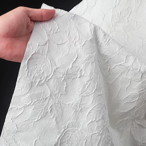 May include: A white fabric featuring a raised, embossed floral pattern with detailed roses and leaves. The material has a textured surface with a matte finish, showing a consistent monochromatic design across the entire piece of textile.