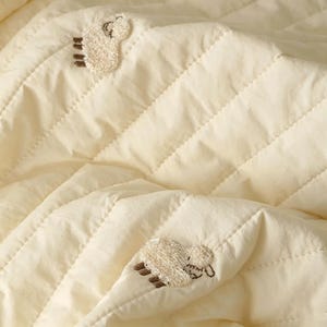 May include: A cream-coloured quilted blanket featuring a repeating pattern of small, embroidered sheep motifs. The fabric has a soft, textured appearance with diagonal stitching lines that create a subtle grid pattern across the surface.