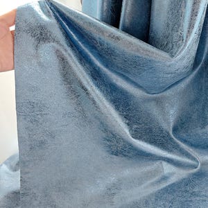 May include: A blue fabric curtain featuring a metallic sheen and a distressed, textured finish. The material displays deep folds and drapes, highlighting the reflective quality of the surface and the uneven, mottled pattern across the blue textile.