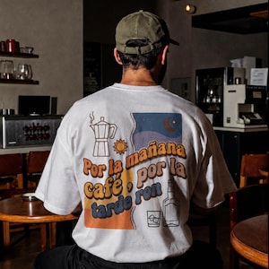 Puede incluir: Una camiseta blanca de manga corta con un gráfico en la espalda que muestra una cafetera italiana, un sol, una luna y una botella con un vaso. El texto dice: Por la mañana café, por la tarde ron. El diseño utiliza tonos naranja, azul y marrón.