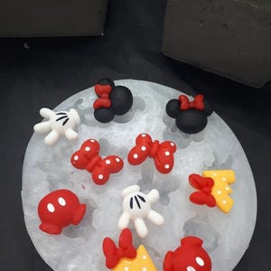 May include: A translucent silicone mould containing various small, colourful character-themed shapes. The shapes include black mouse ears with red bows, white gloves, red polka-dot bows, red shorts with white buttons, and yellow letters with red bows.