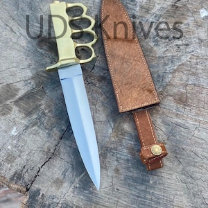 May include: A double-edged dagger with a brass knuckle-duster handle and a silver-toned blade, accompanied by a brown textured leather sheath featuring a brass snap button. The text UDSKnives is visible across the top of the image.