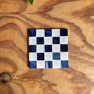 May include: A square coaster featuring a checkerboard pattern made of alternating white and dark blue glass tiles. The tiles have a smooth, glossy finish with subtle variations in the blue color, creating a grid of sixteen individual squares.