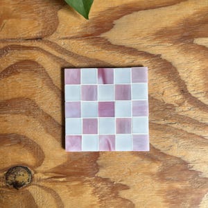 May include: A square coaster featuring a checkerboard pattern made of alternating light pink and white glass tiles. The tiles have a smooth, glossy finish and are arranged in a grid of five by five squares, creating a repeating geometric design.