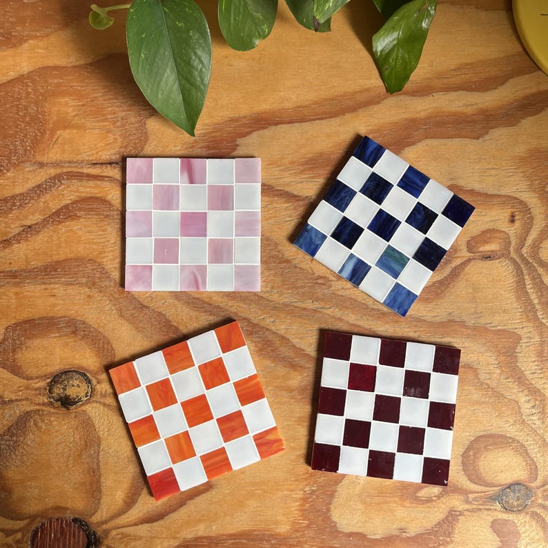 May include: Four square coasters featuring a checkerboard pattern made of small glass tiles. One coaster has pink and white tiles, one has dark blue and white tiles, one has orange and white tiles, and one has deep red and white tiles.