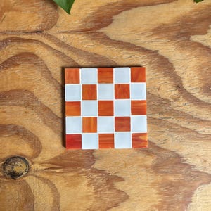 May include: A square coaster featuring a checkerboard pattern made of alternating white and orange glass tiles. The orange tiles have a marbled, semi-translucent appearance, while the white tiles are opaque. The coaster has a flat, smooth surface.