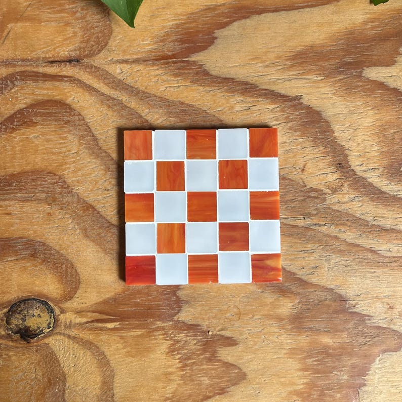 May include: A square coaster featuring a checkerboard pattern made of alternating white and orange glass tiles. The orange tiles have a marbled, semi-translucent appearance, while the white tiles are opaque. The coaster has a flat, smooth surface.