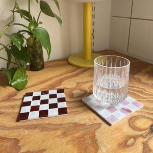 May include: Two square coasters featuring a grid of small tiles. One coaster displays a checkerboard pattern of deep red and white tiles. The second coaster features a grid of light pink and white tiles, with a clear, vertically ribbed glass tumbler.