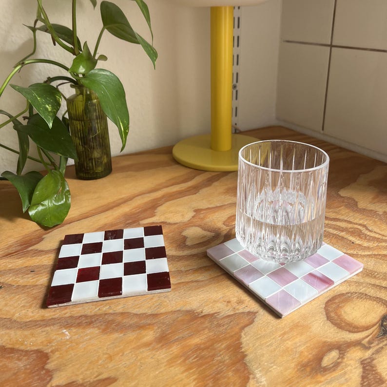 May include: Two square coasters featuring a grid of small tiles. One coaster displays a checkerboard pattern of deep red and white tiles. The second coaster features a grid of light pink and white tiles, with a clear, vertically ribbed glass tumbler.
