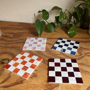 May include: Four square coasters featuring a checkerboard pattern. One coaster has pink and white tiles, one has dark blue and white tiles, one has orange and white tiles, and one has deep red and white tiles. Each coaster is made of small glass squares.