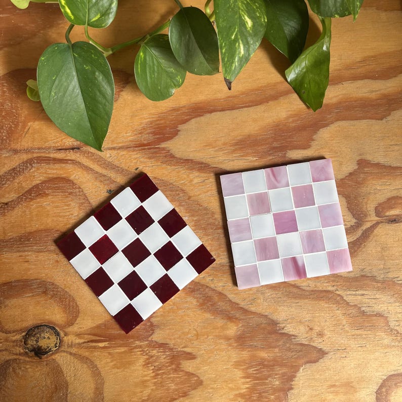 May include: Two square coasters featuring a checkerboard pattern. One coaster displays alternating deep burgundy and white tiles, while the other features alternating light pink and white tiles. Both coasters are arranged on a wooden surface.