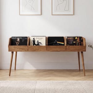 Può includere: Un mobile in legno per riporre dischi in vinile, dotato di quattro scomparti distinti per organizzare gli album. L'unità poggia su quattro gambe affusolate e contiene diversi dischi, tra cui copertine di Miles Davis, Fleetwood Mac e Pink Floyd.