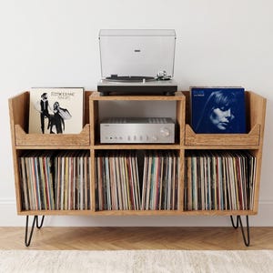 Puede incluir: Un mueble de madera de color marrón claro para almacenar discos de vinilo con patas de horquilla de metal negro. La unidad cuenta con un tocadiscos superior, un amplificador central y dos compartimentos laterales con discos, incluyendo Fleetwood Mac Rumours.