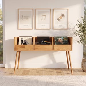 Può includere: Un mobile in legno chiaro per riporre dischi in vinile con tre scomparti aperti, ognuno contenente un album. Il mobile è dotato di quattro gambe affusolate ed è posizionato contro una parete bianca con tre stampe artistiche a tema musicale.