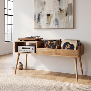 May include: A light oak wood media console featuring four tapered legs, an open shelf housing a silver audio receiver, and two integrated storage compartments filled with vinyl records, with a turntable placed on the top surface.