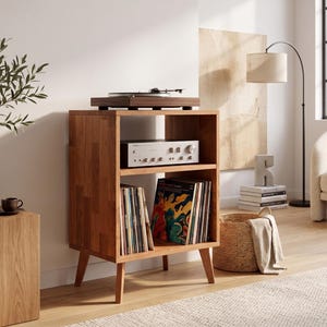 May include: A wooden record player stand with four tapered legs, featuring an open top surface holding a turntable, a middle shelf with a silver audio receiver, and a bottom shelf filled with vinyl records organized in vertical rows.
