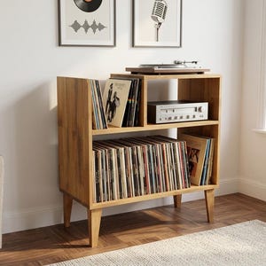 Puede incluir: Un mueble de madera de color marrón claro para almacenar discos con cuatro patas cónicas. La unidad cuenta con un estante superior abierto dividido en dos secciones para discos de vinilo y equipo de audio, y un estante inferior lleno de vinilos.