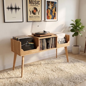 May include: A light wood vinyl record storage console with tapered legs, featuring three open compartments filled with vinyl records and a turntable with a clear dust cover placed on the centre top surface of the wooden furniture unit.