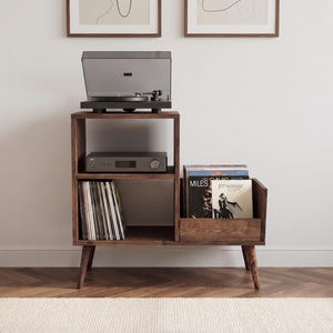 May include: A dark brown wooden record player stand with four tapered legs. The unit features an open top shelf holding a black turntable, a middle shelf with a grey audio receiver, and a lower section with vertical vinyl record storage.