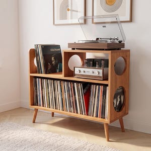 May include: A wooden record player console with tapered legs, featuring a top surface for a turntable and amplifier, and two lower shelves filled with vinyl records. The unit has circular cutouts on the side panels and a warm brown wood finish.
