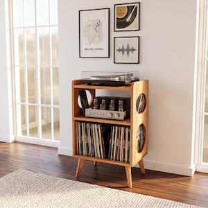 Può includere: Un mobiletto per giradischi in legno chiaro con quattro gambe affusolate, dotato di un ripiano superiore per il giradischi, un ripiano centrale con un amplificatore e un ripiano inferiore pieno di dischi in vinile. I pannelli laterali hanno aperture circolari.