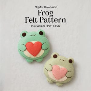 May include: Digital download Frog Felt Pattern with instructions, PDF, and SVG. Two felt frog shapes are shown, one light green with a red heart and one pale green with a pink heart, both featuring dark brown eyes and stitched details.