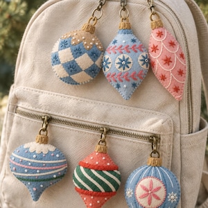 May include: A beige canvas backpack adorned with six felt ornaments attached to the zippers. The ornaments feature various shapes and embroidered patterns in blue, white, pink, red, and green, each finished with a small gold-toned metal cap and clasp.