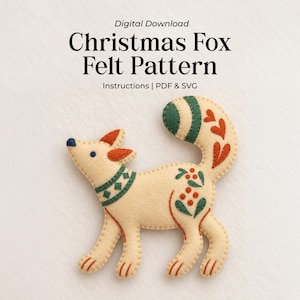May include: A digital download for a Christmas Fox Felt Pattern including PDF and SVG instructions. The image shows a cream-coloured felt fox ornament with dark green and orange embroidered details, including a patterned collar and tail.