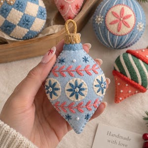 May include: A collection of teardrop and round felt Christmas ornaments featuring intricate embroidery in blue, pink, green, and cream. Each ornament is decorated with stitched floral patterns, small white beads, and a gold-colored hanging loop.