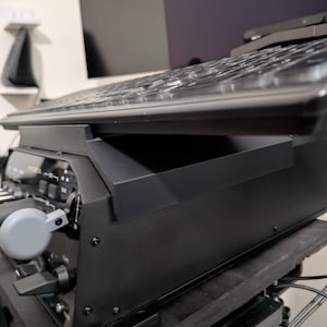 Op de afbeelding: Een zwart bedieningspaneel voor een vluchtsimulator met een horizontaal toetsenbord aan de bovenzijde. Het apparaat bevat een grijze ronde knop, diverse zwarte tuimelschakelaars en witte tekstlabels op de schuine voorzijde van de unit.