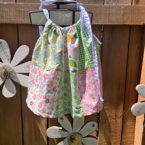 May include: A sleeveless dress featuring a patchwork design of various floral and fruit-patterned fabrics in shades of pink, green, yellow, and white. The dress has a gathered neckline with white fabric ties at the shoulders and a loose, flared fit.