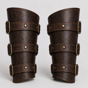 Puede incluir: Un par de protectores de brazo texturizados en color marrón oscuro con tres correas horizontales, hebillas de metal en tono latón antiguo y remaches a juego. Los protectores tienen una forma curva y cónica con un grano visible.