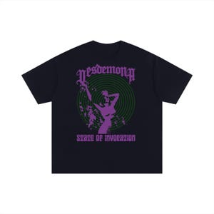以下が含まれることがあります： 黒の半袖Tシャツ。濃い緑色の渦巻き模様を背景に、腕を上げた人物の紫色のグラフィックが描かれています。上部にはゴシック体で「Desdemona」、下部には紫色の文字で「State of Invocation」とプリントされています。