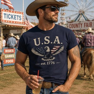 May include: A navy blue short-sleeve t-shirt featuring a graphic of a bald eagle with wings spread, partially colored like the American flag. The text U.S.A. is printed above the eagle, and the text est. 1776 is printed below the eagle in white.