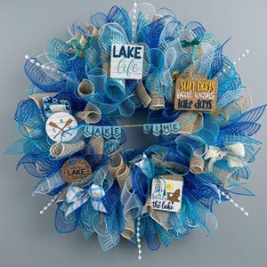 May include: A decorative wreath featuring blue and tan mesh ribbons, white beaded accents, and wooden signs with text including Lake Life, Sun Days, Lake Days, Lake Time, and Beach at the Lake, along with a small wooden cutout of two crossed oars.