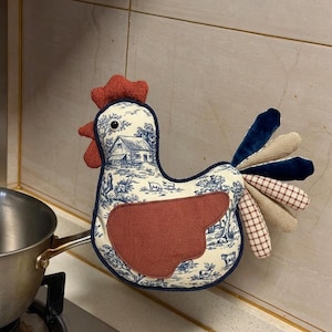 Op de afbeelding: Een stoffen ovenwant in de vorm van een kip met een blauw-wit toile de Jouy-patroon van een boerderijtafereel. De want heeft een roestkleurige vleugel, een getextureerde rode kam en lellen, en een staart van fluwelen en geruite stoffen stroken.