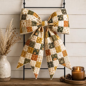 May include: A large decorative bow made of quilted fabric featuring a patchwork pattern of squares in shades of olive green, mustard yellow, orange, and cream. The fabric displays various prints including small pumpkins, floral motifs, and plaid.