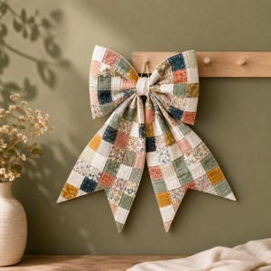 May include: A large decorative bow constructed from quilted fabric featuring a patchwork pattern of squares in muted tones of sage green, mustard yellow, navy blue, and cream, with some squares displaying small floral prints and gingham checks.