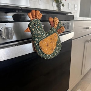 May include: A chicken-shaped oven mitt featuring a dark green fabric body with a white acorn and leaf print, a mustard yellow floral wing patch, a solid orange comb and beak, and a multi-colored plaid tail section hanging from a stainless steel oven handle.