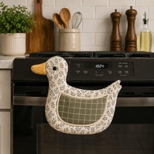 May include: A duck-shaped oven mitt featuring a cream-colored fabric with a small black botanical print. The duck has a yellow beak, a black eye, and a green wing patch with a white grid pattern. The mitt is hanging on the handle of a black oven.